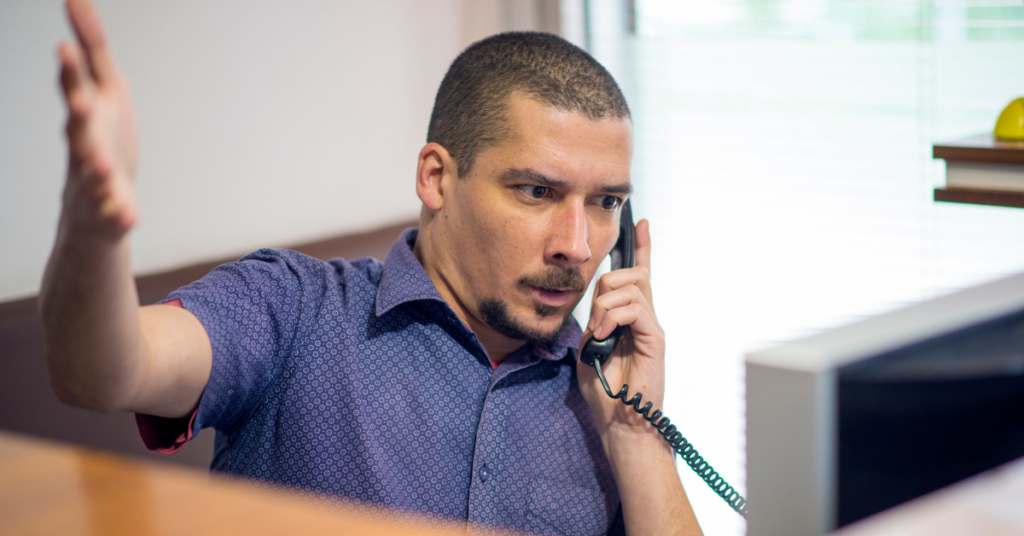 Man on phone at desk looking concerned with arm in the air.