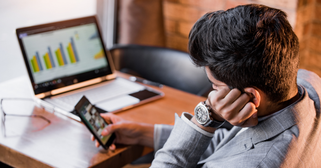 male scrolling on phone procrastinating while laptop open on desk.